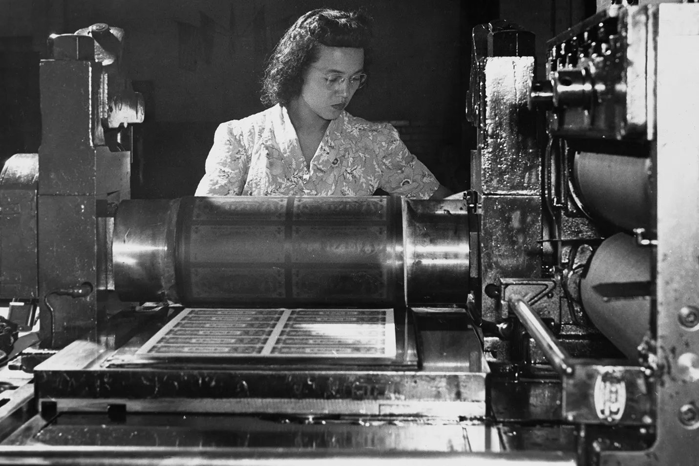 A historic photo of a woman working with a large printing press to print currency.